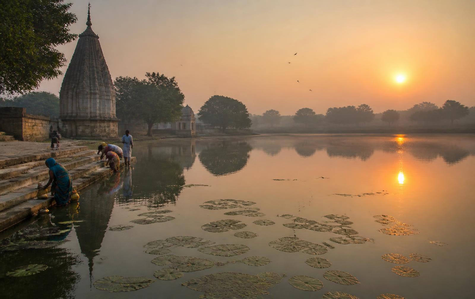 A large temple tank at sunrise with lotus leaves, stone bathing steps, villagers filling brass pots, and a temple spire reflected in the water