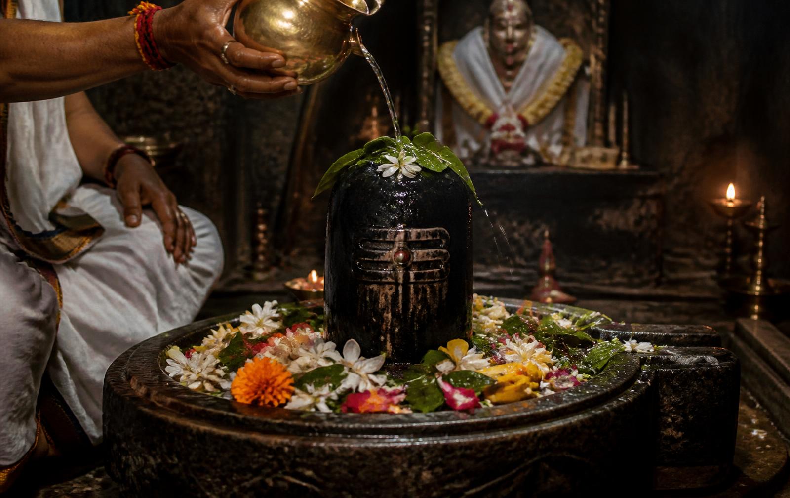 Water being poured from a brass vessel over a black stone Shiva lingam covered in bel leaves and flowers inside a dim sanctum