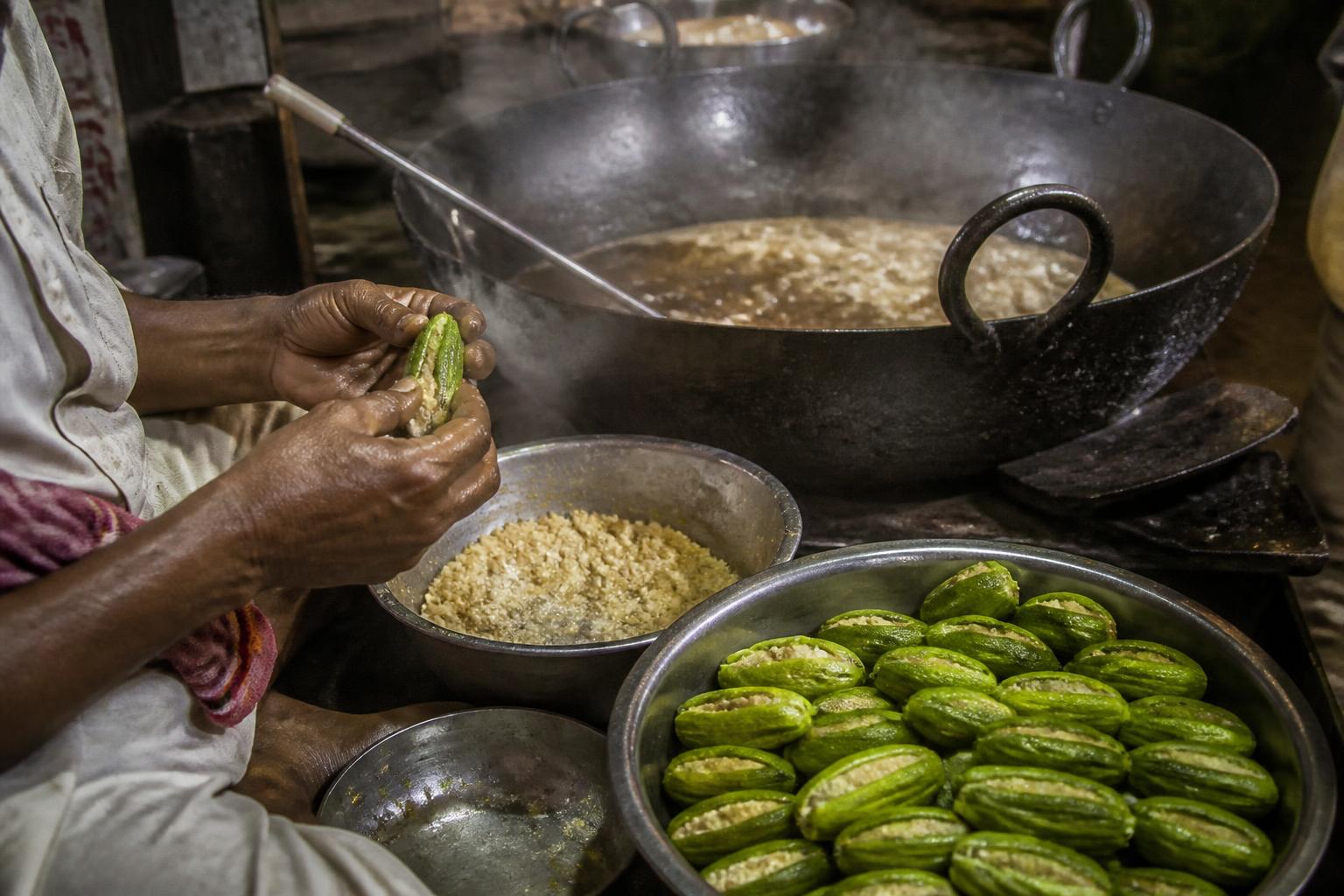 A halwai scooping seeds from blanched pointed gourds and filling them with khoya beside a steaming kadhai of syrup